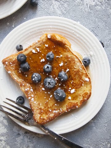 Crispy challah French toast with cinnamon on a plate with maple syrup, blueberries, and powdered sugar with a fork and napkin.