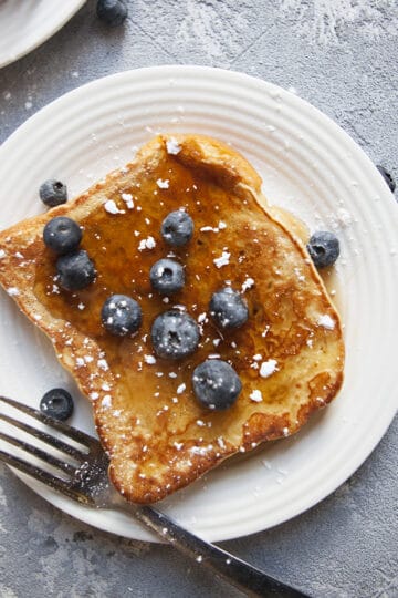 Crispy challah French toast with cinnamon on a plate with maple syrup, blueberries, and powdered sugar with a fork and napkin.