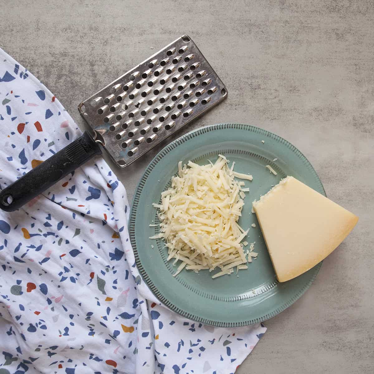 Wedge of Parmesan cheese next to grated Parmesan on a blue plate alongside a cheese grater and a terrazzo-patterned kitchen towel on a gray background.