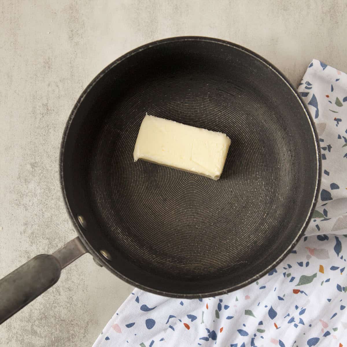 A stick of butter in a black saucepan on a gray background with a terrazzo-patterned kitchen towel.