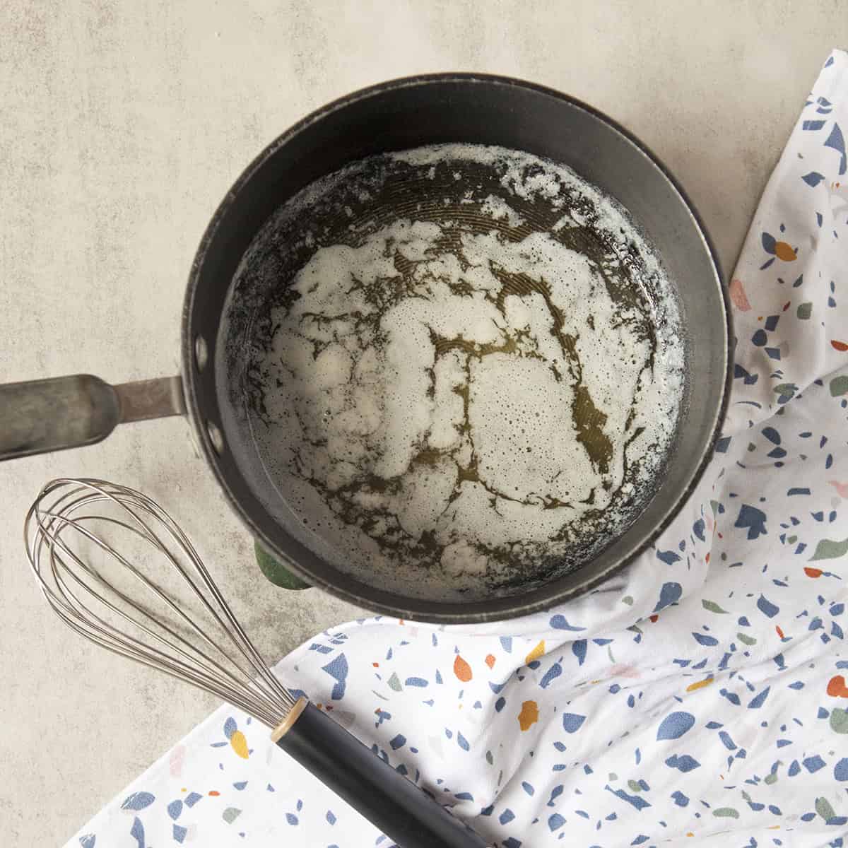 Melted butter in a black saucepan next to a whisk and a terrazzo-patterned kitchen towel on a gray background.