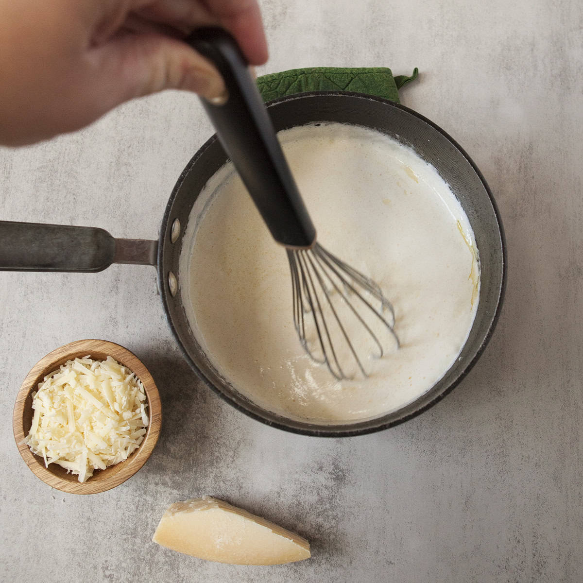 Heavy cream being whisked in a black saucepan nexto to a small bowl of grated Parmesan and a small wedge of Parmesan cheese on a gray background.