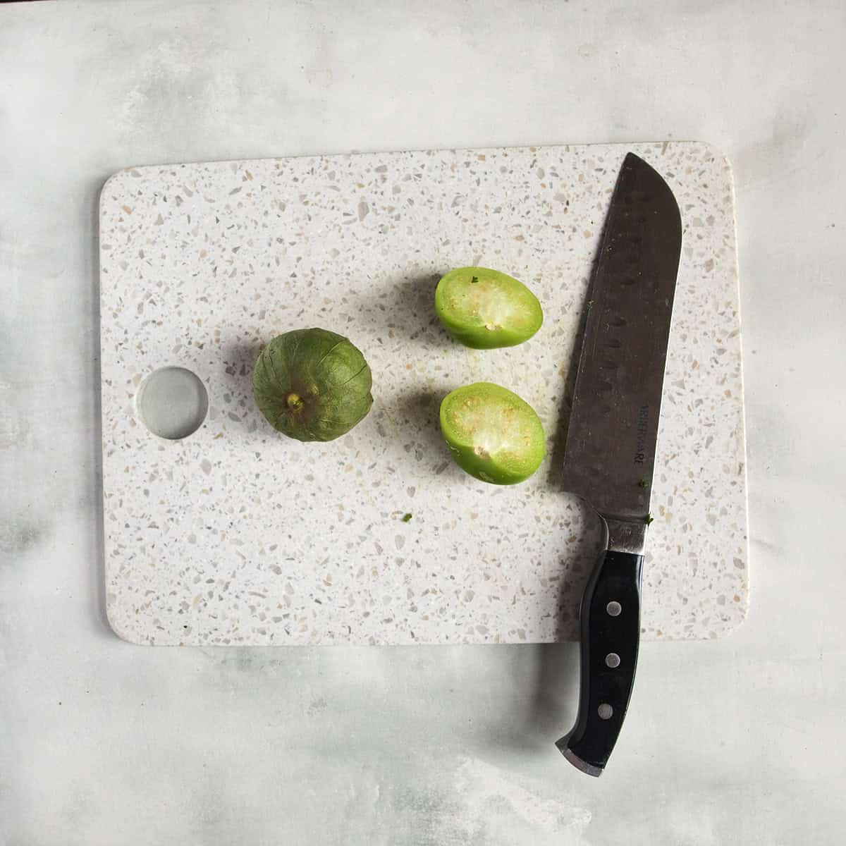 A tomatillo chopped in half alongside a whole tomatillo on a cutting board next to a knife on a gray background.