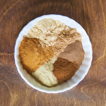 Chai spice ingredients (ground cinnamon, ginger, cardamom, nutmeg, cloves, and allspice) in a bowl on a wooden backdrop