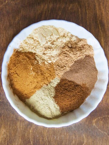 Chai spice ingredients (ground cinnamon, ginger, cardamom, nutmeg, cloves, and allspice) in a bowl on a wooden backdrop