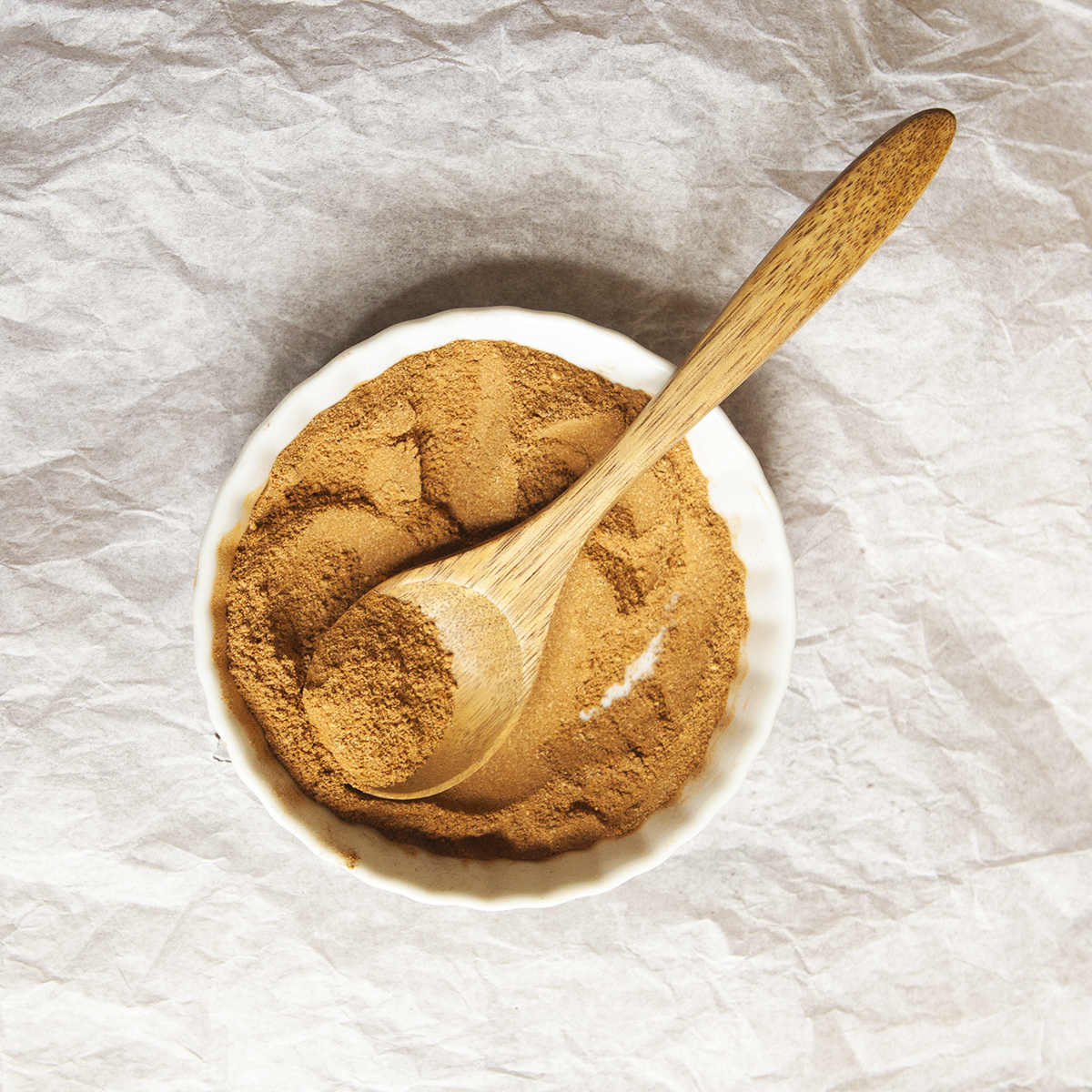 Ground chai spice blend in a small bowl with a wooden spoon on a white tissue paper backdrop.