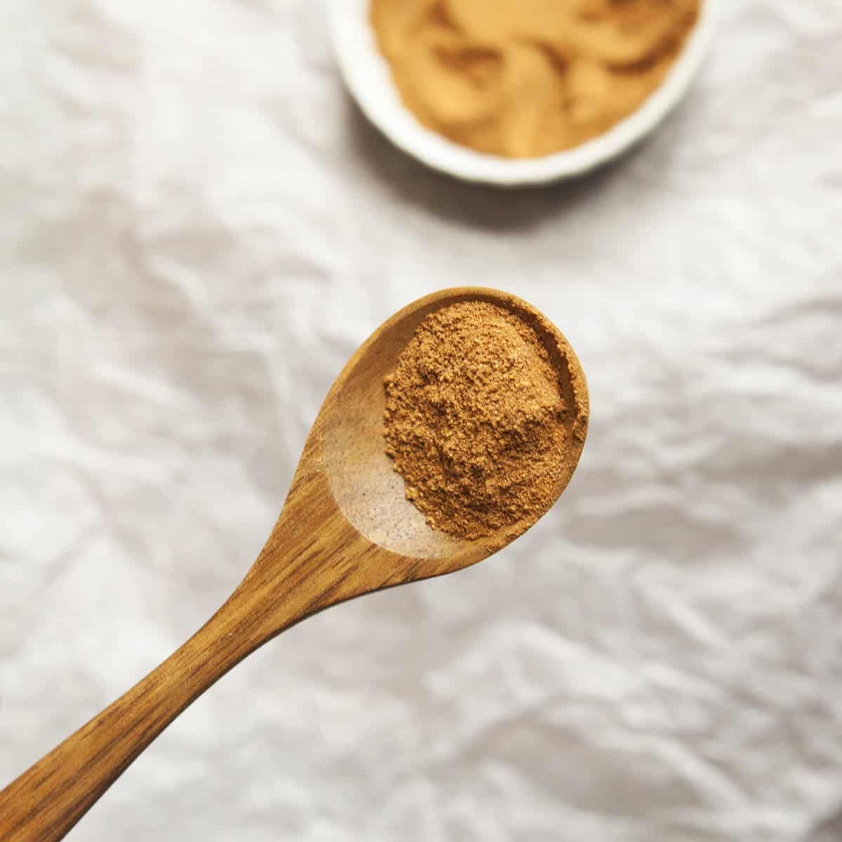 Orange-tan colored powdered chai spice blend on a wooden spoon above a white tablecloth next to a small bowl of chai spice blend.