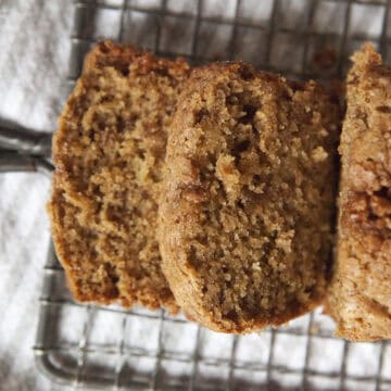 Golden brown homemade chai-spiced banana bread loaf with two slices resting on a wire grater on top of a white dish towel.