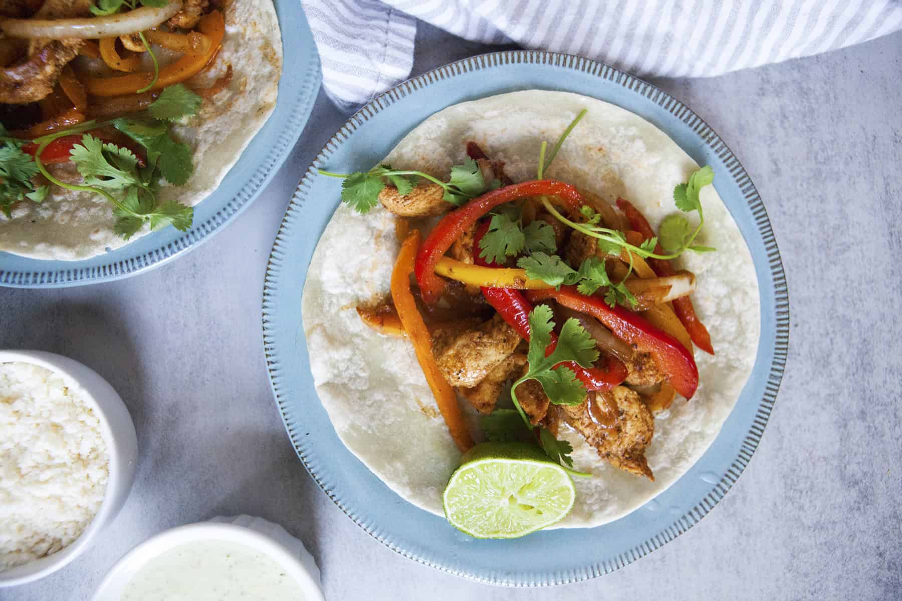 Chicken fajitas with onions and bell peppers on a flour tortilla, sitting on a blue plate with half a lime, on a gray surface with grated cheese, a napkin, and jalapeno ranch sauce.