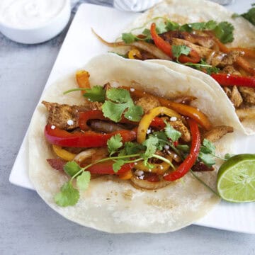 Fajitas with sliced chicken, bell peppers, onions, cilantro, and cheese on a flour tortilla, sitting on a square white plate with a sliced lime.