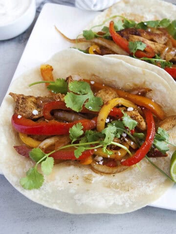 Skillet chicken fajitas with seasoned chicken strips, bell peppers, onions, cilantro, lime juice, and Cotija cheese on a flour tortilla with a sliced lime on a square white plate.