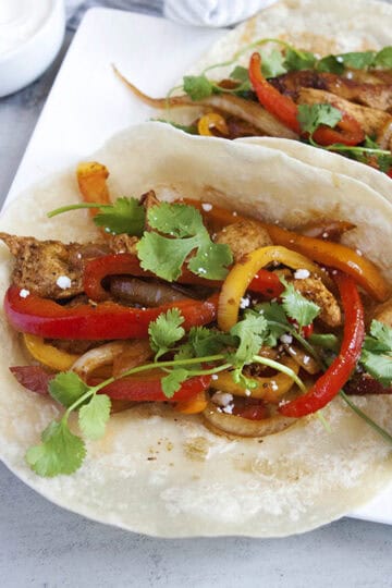 Skillet chicken fajitas with seasoned chicken strips, bell peppers, onions, cilantro, lime juice, and Cotija cheese on a flour tortilla with a sliced lime on a square white plate.