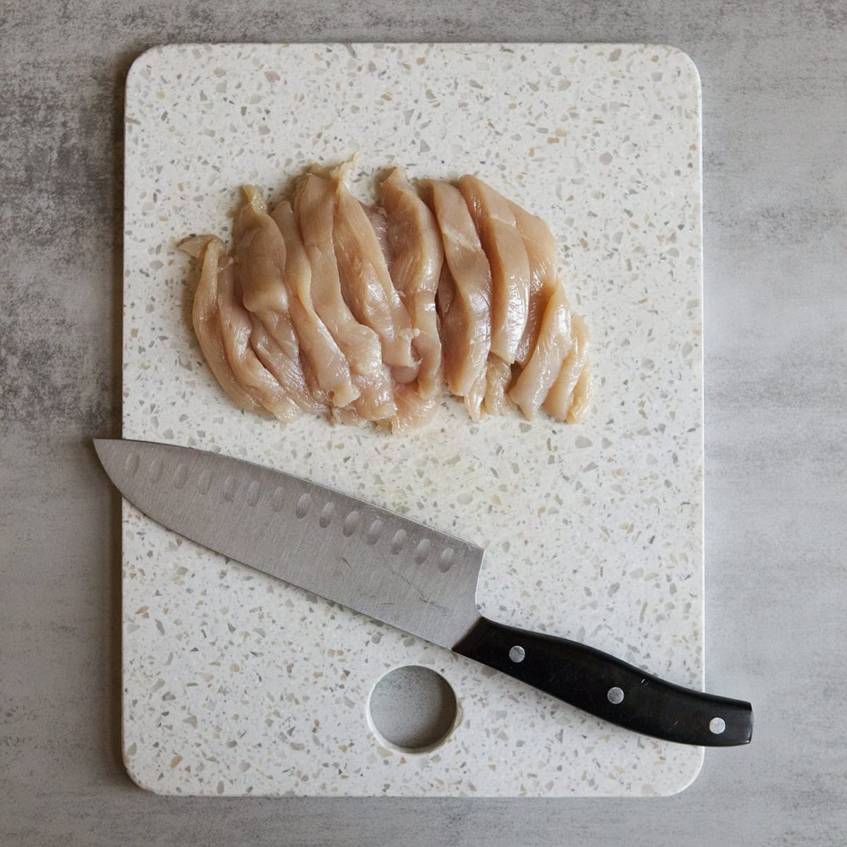 Strips of raw chicken breast on a cutting board with a knife.