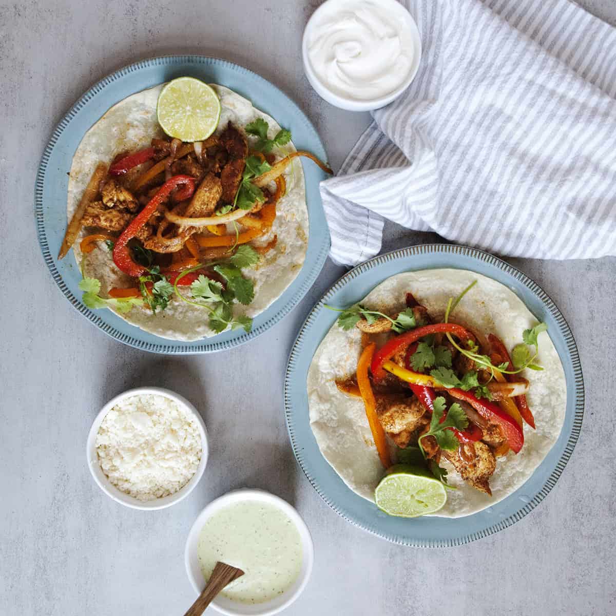 chicken fajitas with sliced onion, bell pepper, cilantro, and Cotija on flour tortillas, sitting on 2 blue plates on a gray background.  Nearby are a napkin, sour cream, Cotija cheese, and creamy jalapeno ranch dressing in small bowls.