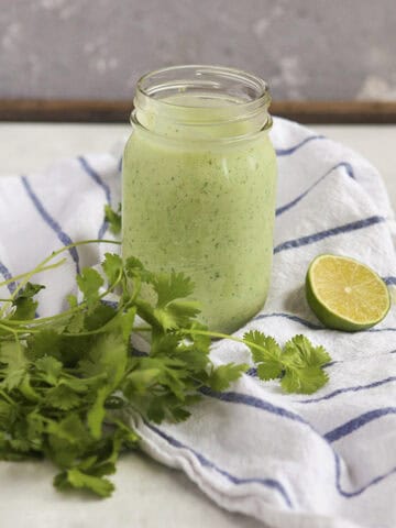 Creamy jalapeno tomatillo ranch dressing with cilantro (Cafe Rio copycat) in a Mason jar sitting on a blue and white striped dish towel next to a bunch of cilantro and half of a lime on a gray background.