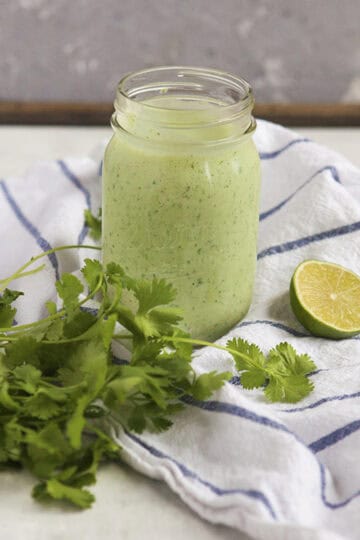 Creamy jalapeno tomatillo ranch dressing with cilantro (Cafe Rio copycat) in a Mason jar sitting on a blue and white striped dish towel next to a bunch of cilantro and half of a lime on a gray background.