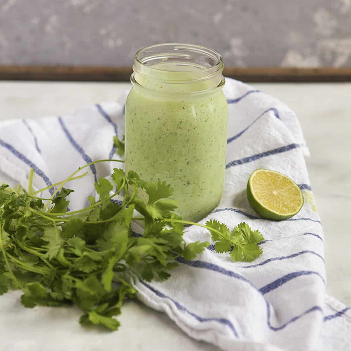 Creamy jalapeno tomatillo ranch dressing with cilantro (Cafe Rio copycat) in a Mason jar sitting on a blue and white striped dish towel next to a bunch of cilantro and half of a lime on a gray background.