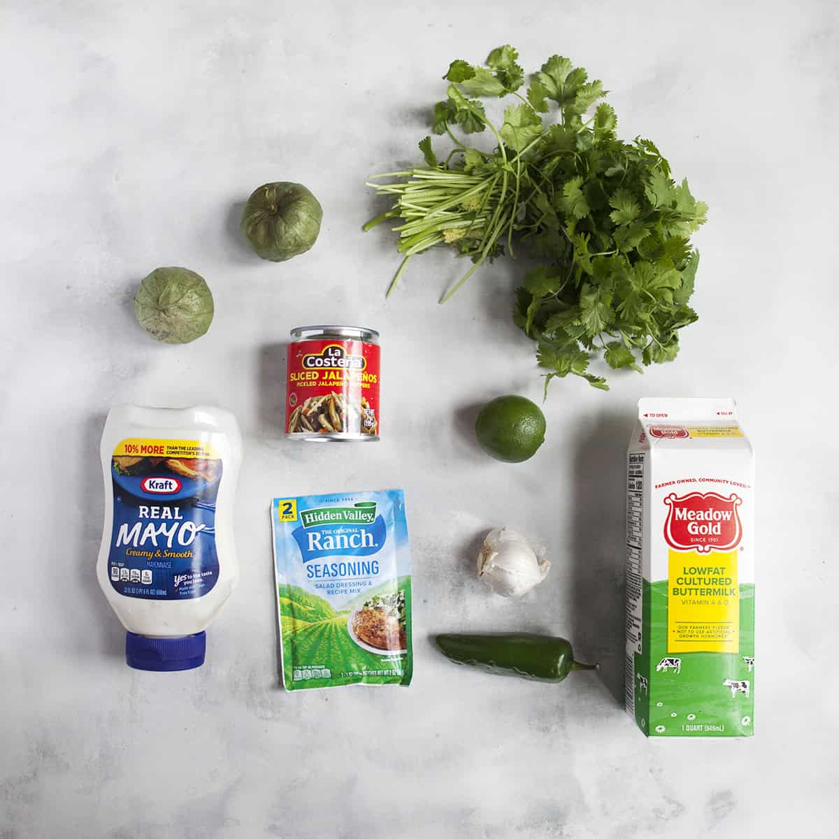 Ingredients for creamy jalapeno ranch dressing with cilantro:  mayonnaise, Hidden Valley Ranch seasoning packet, buttermilk, jalapeno, lime, tomatillo, garlic, pickled jalapenos, cilantro, on a gray backdrop.