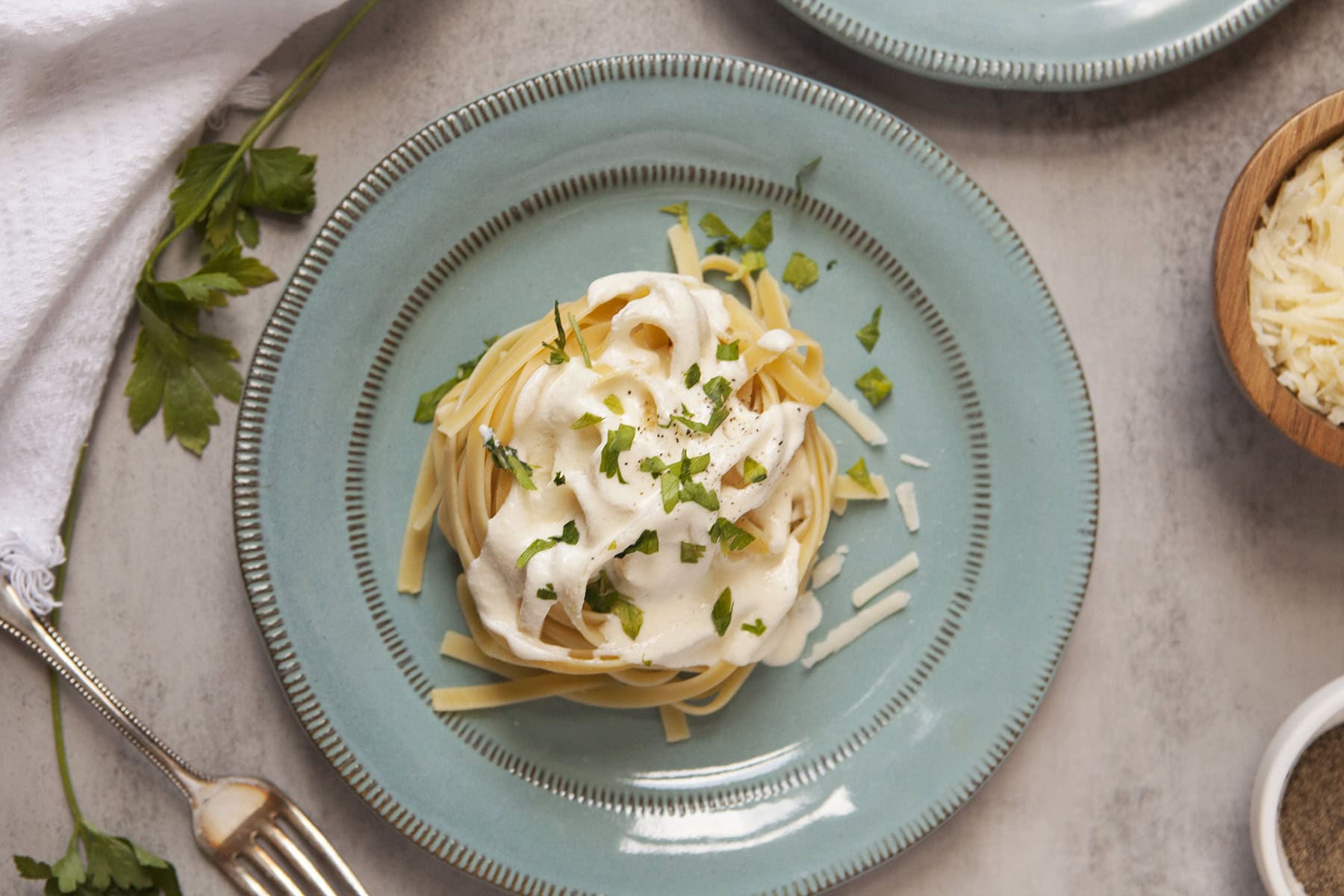 Fettuccine Alfredo nest with chopped parsley on a blue plate on a gray background with a fork, sprig of parsley, white napkin, and bowls with shredded Parmesan and pepper.