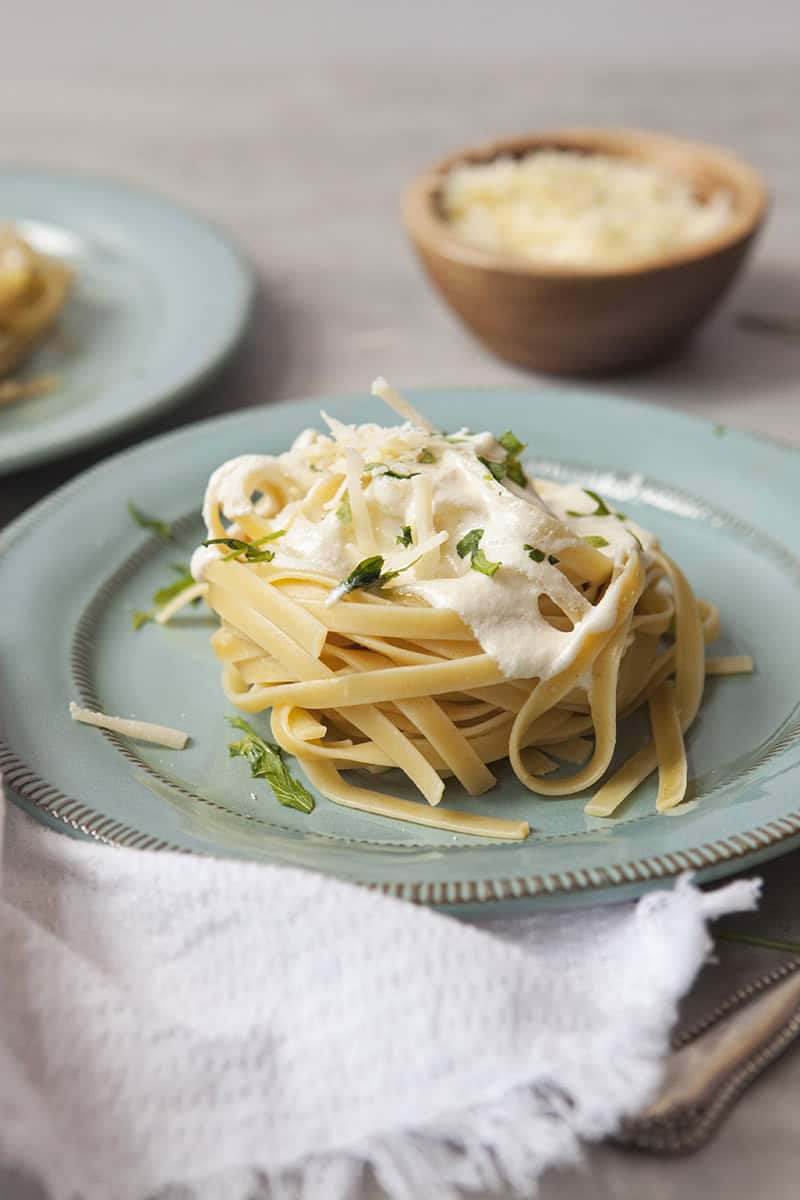 Fettuccine alfredo nest on a blue plate, topped with chopped parsley, on a table with a second plate, small wooden bowl with grated Parmesan cheese, and a white fringed napkin.