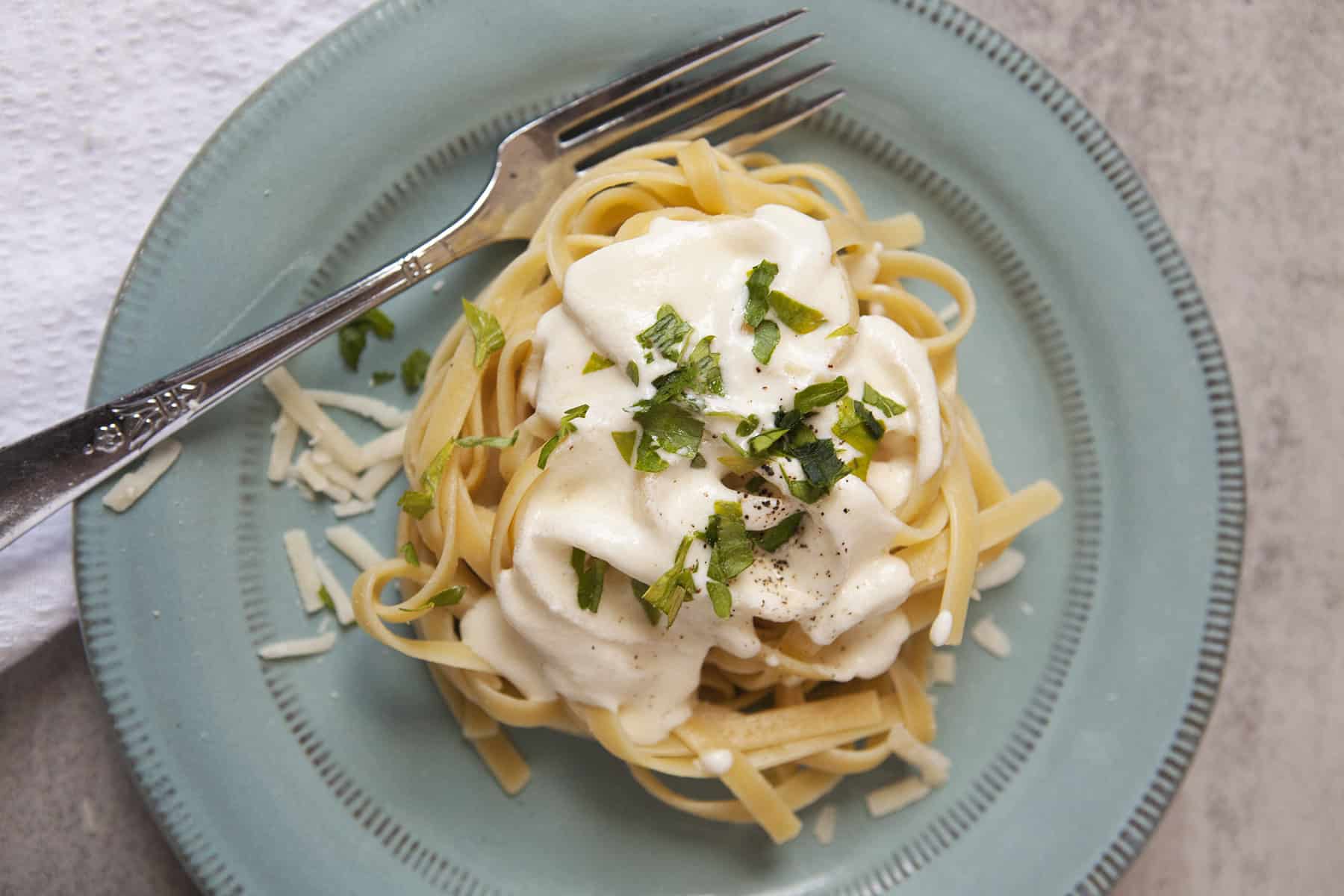 Fettuccine Alfredo nest with chopped parsley and pepper on a blue plate with a fork, on a gray background with a white napkin.