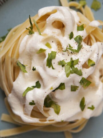 Thick, creamy Alfredo sauce on a nest of fettuccine, topped with chopped parsley and pepper, on a blue plate.