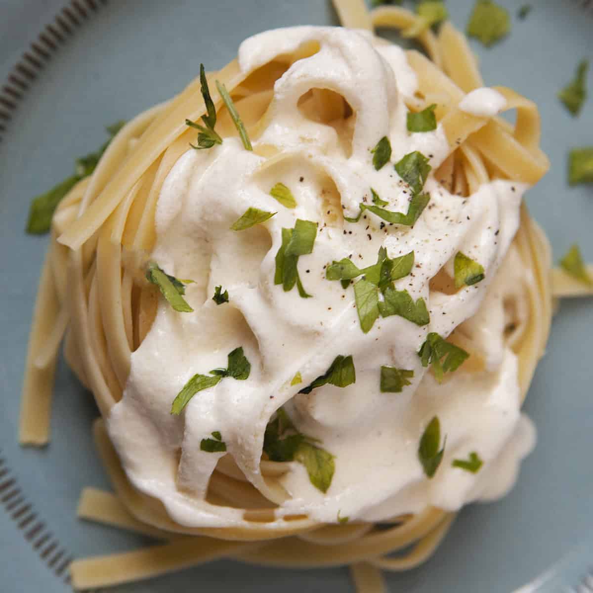 Thick, creamy Alfredo sauce on a nest of fettuccine, topped with chopped parsley and pepper, on a blue plate.