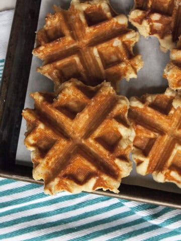 Crispy brown Belgian Liege waffles on a baking sheet on top of a turquoise and white striped kitchen towel.