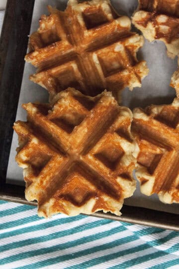 Crispy brown Belgian Liege waffles on a baking sheet on top of a turquoise and white striped kitchen towel.