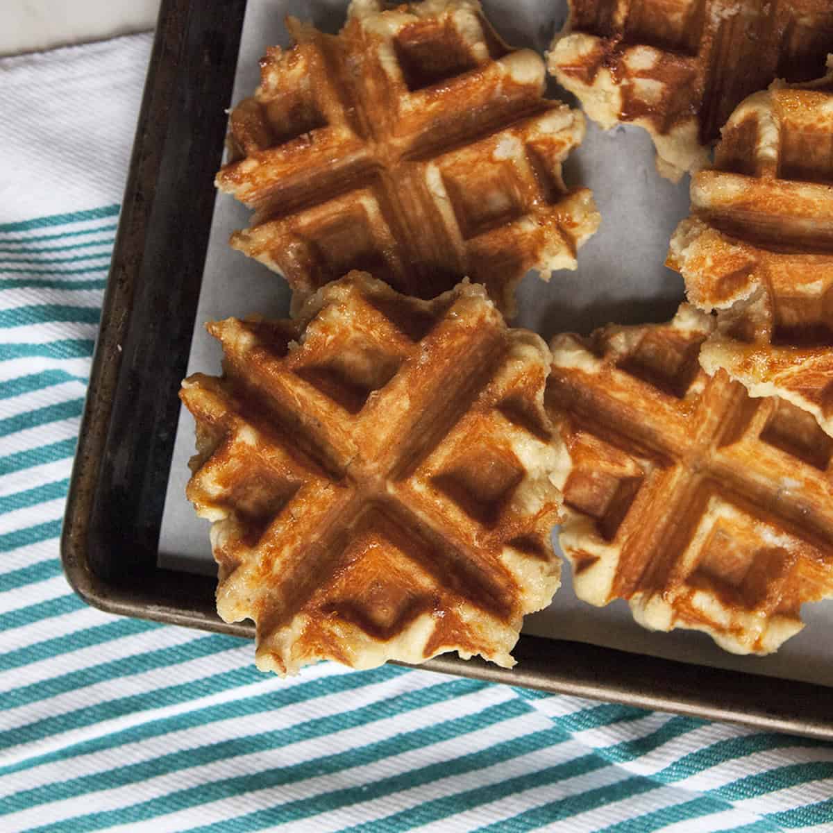 Crispy brown Belgian Liege waffles on a baking sheet on top of a turquoise and white striped kitchen towel.