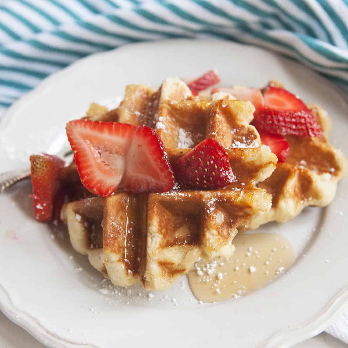 Golden brown Belgian Liege waffles on a white plate topped with sliced strawberries, powdered sugar, and maple syrup next to a turquoise and white striped kitchen towel.