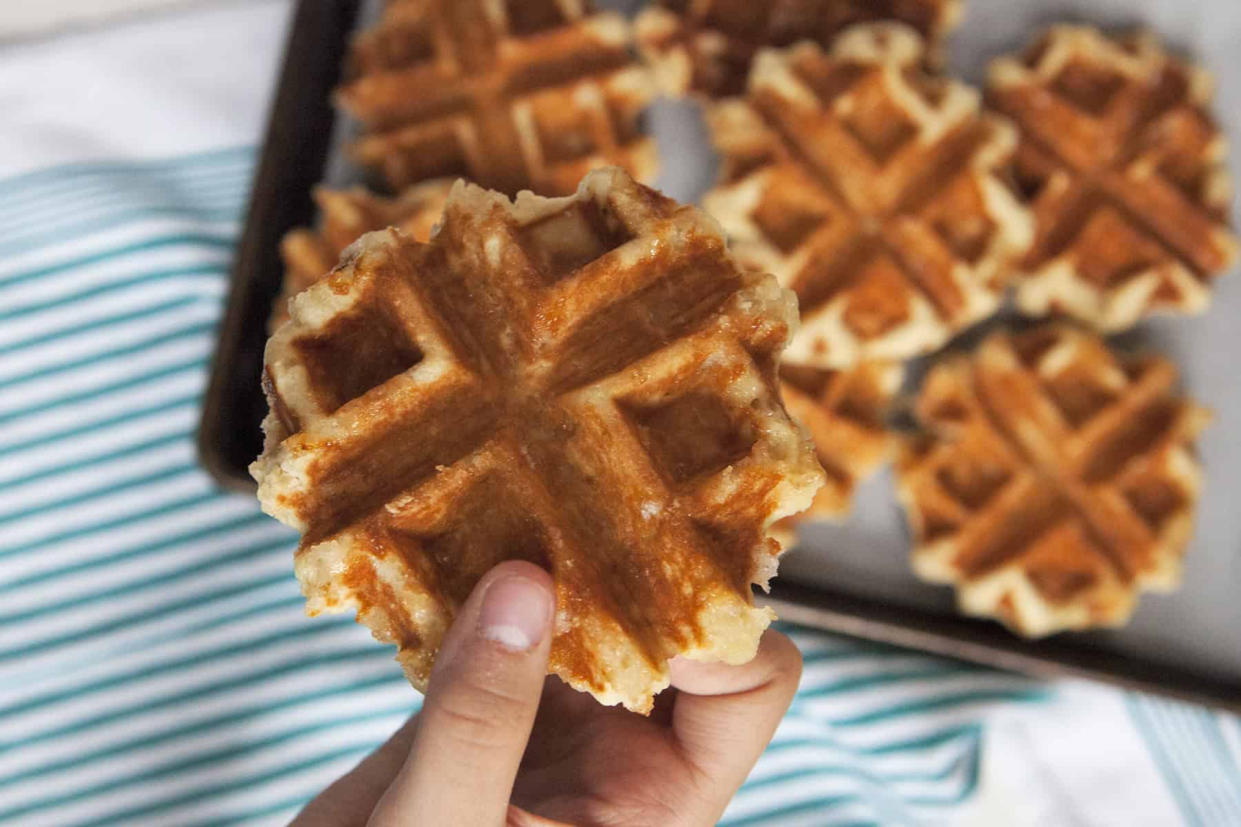 A hand holding a golden brown Belgian Liege waffle over a baking tray filled with more waffles, next to a turquoise and white striped kitchen towel.