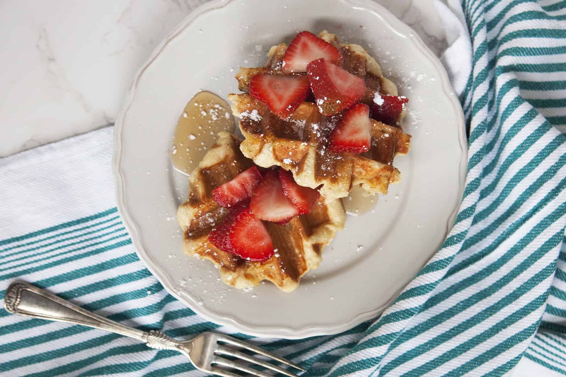 Golden brown Belgian Liege waffles on a white plate topped with sliced strawberries, maple syrup, and powdered sugar, on a marble countertop next to a fork and a turquoise and white striped kitchen towel.