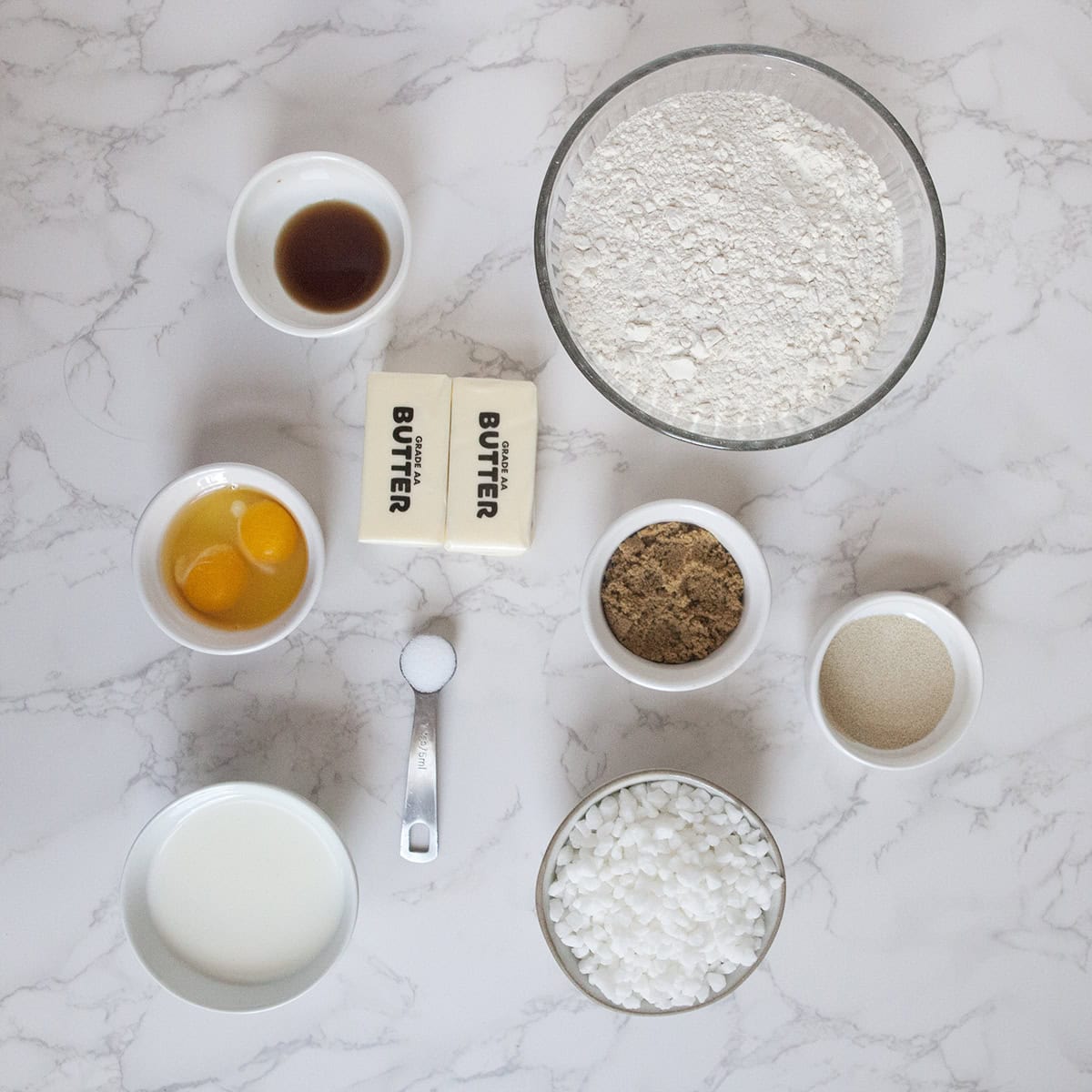 Ingredients for Belgian Liege waffles displayed on a marble countertop:  AP flour, milk, yeast, brown sugar, salt, eggs, butter, vanilla, and pear sugar, all in individual small bowls.