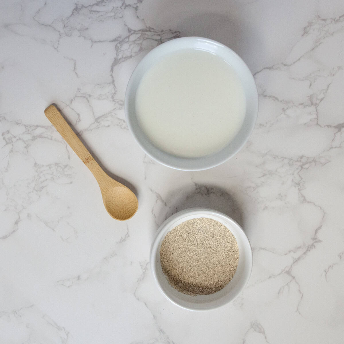 Small bowls containing milk and active dry yeast on a white marble countertop next to a small wooden spoon.