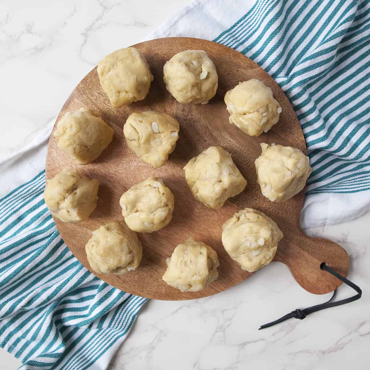 Twelve balls of Belgian Liege waffle dough on a serving board on a white marble countertop with a turquoise and white striped kitchen towel.