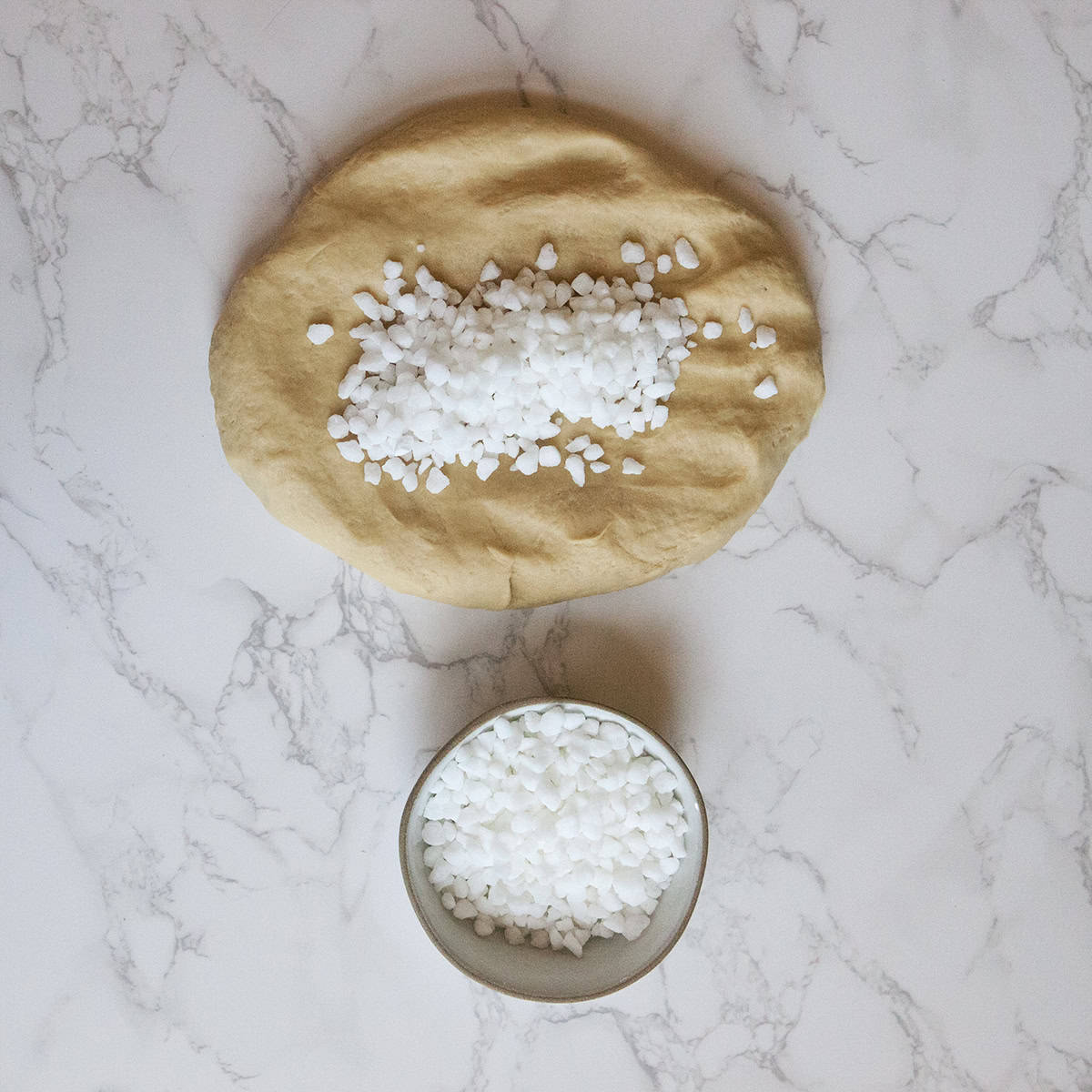 Belgian Liege waffle dough on a white marble countertop topped with Belgian pearl sugar, with more Belgian pearl sugar in a small bowl.