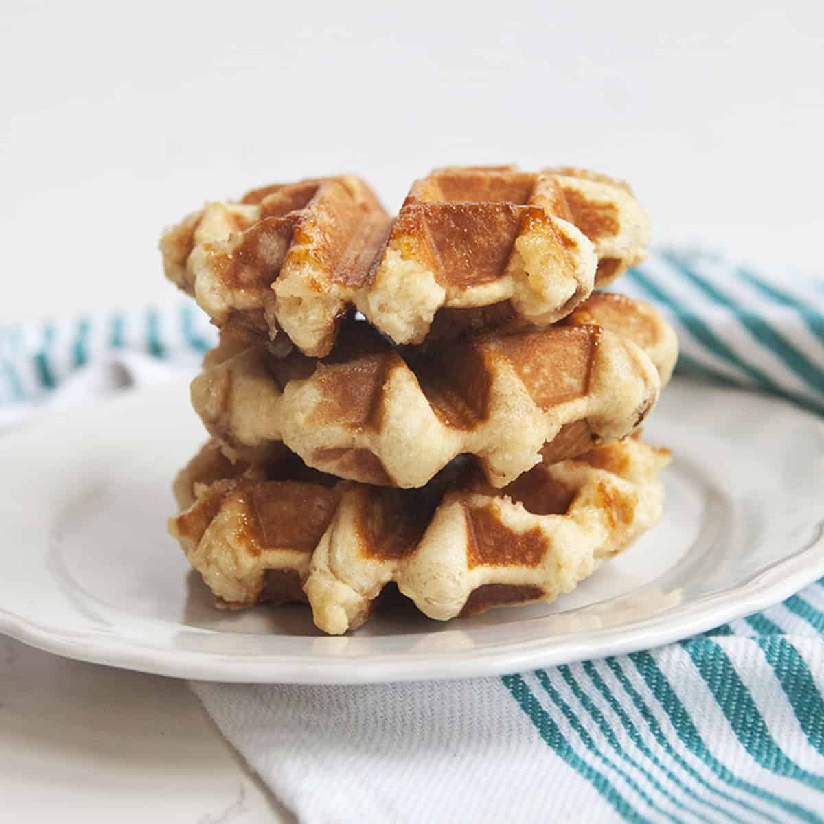 Three golden brown Belgian Liege waffles stacked on a white plate on top of a turquoise and white striped dishcloth with a white backdrop.