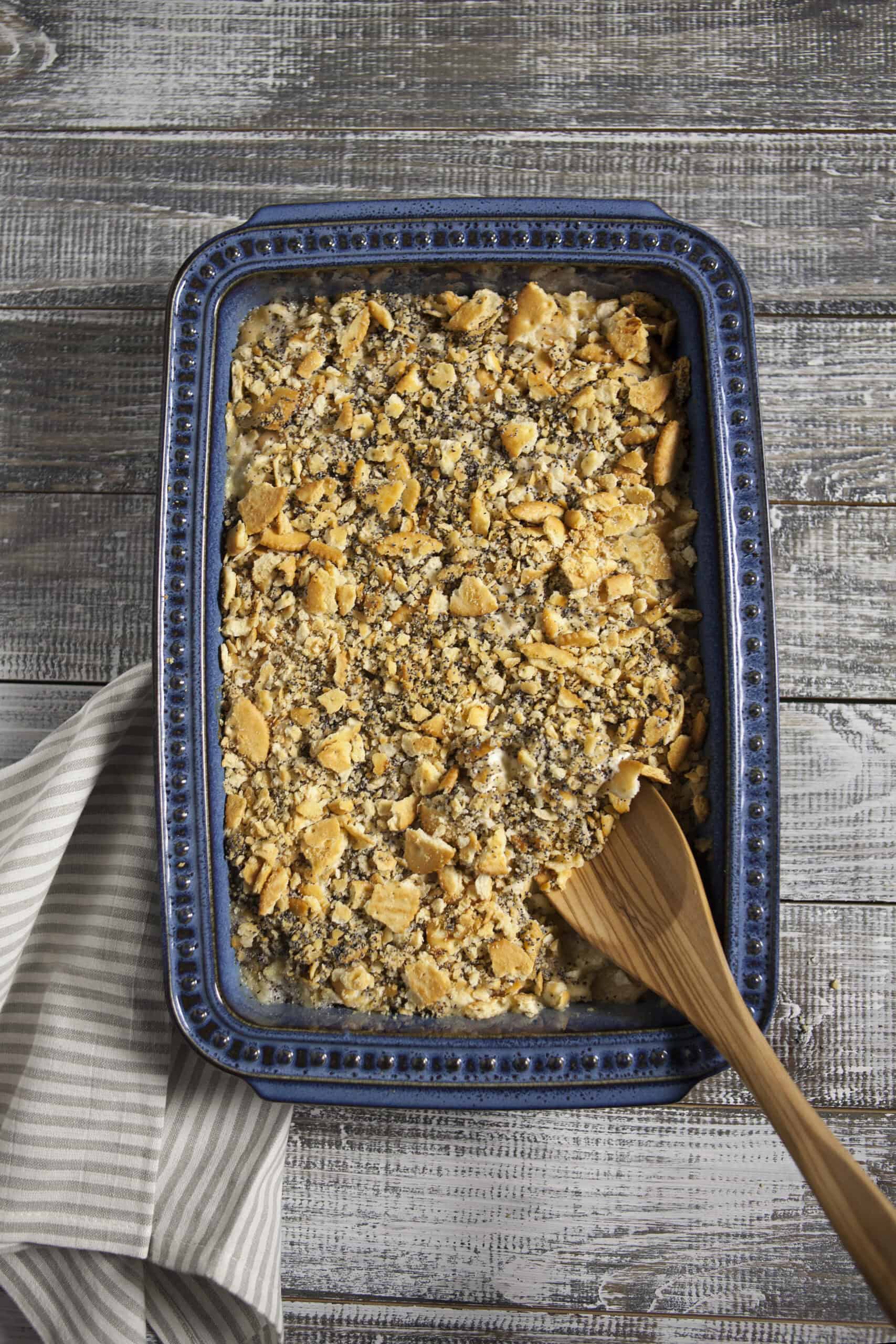 Cooked poppy seed chicken casserole with a topping of crushed Ritz buttery crackers and poppy seeds in a blue 9x13-inch baking dish with a wooden spoon next to a white and gray striped kitchen towel on a wooden table.