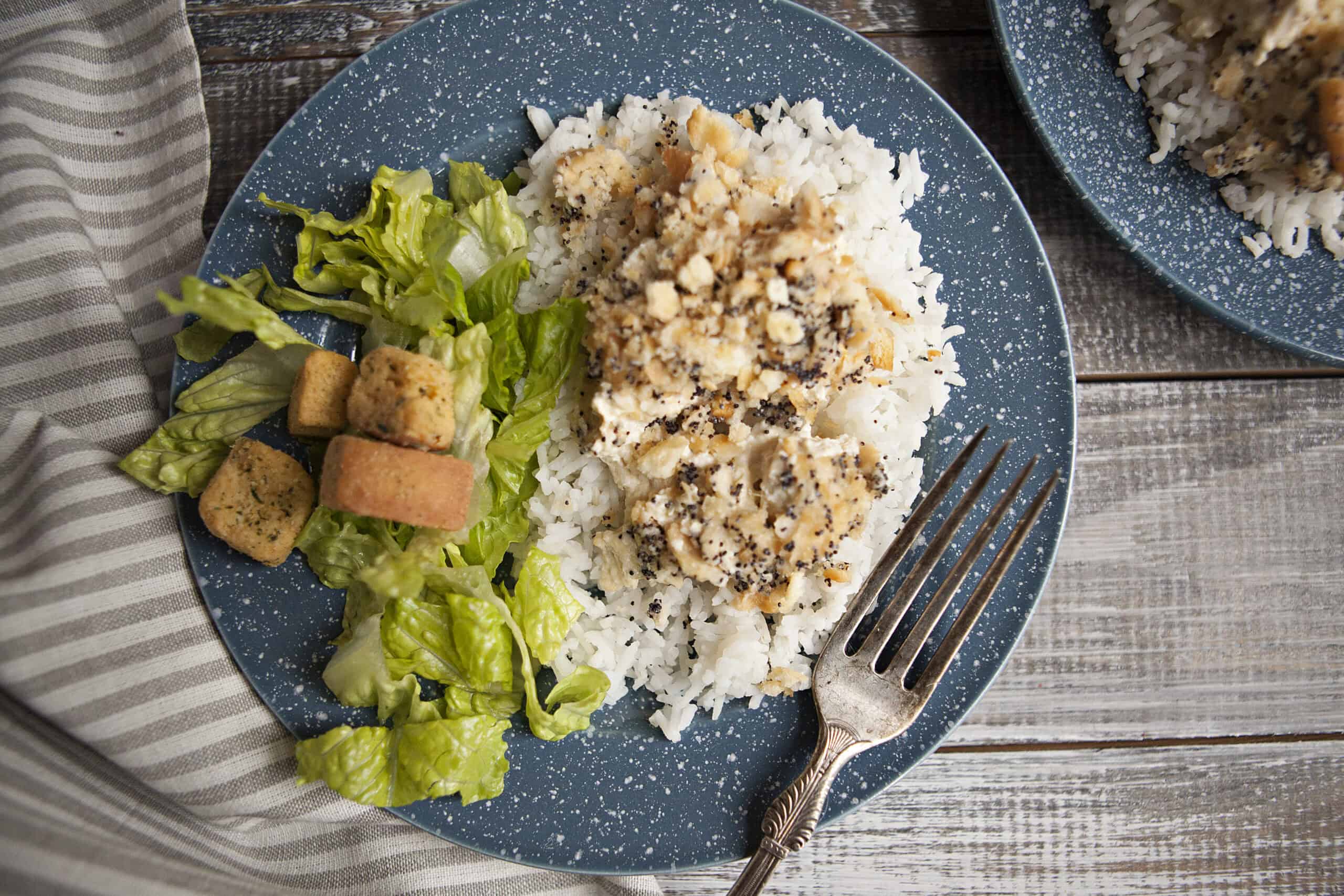 Poppy seed chicken casserole on white rice next to a green salad with croutons on a blue plate with white speckles on a wooden table next to a fork and a white and gray striped napkin.