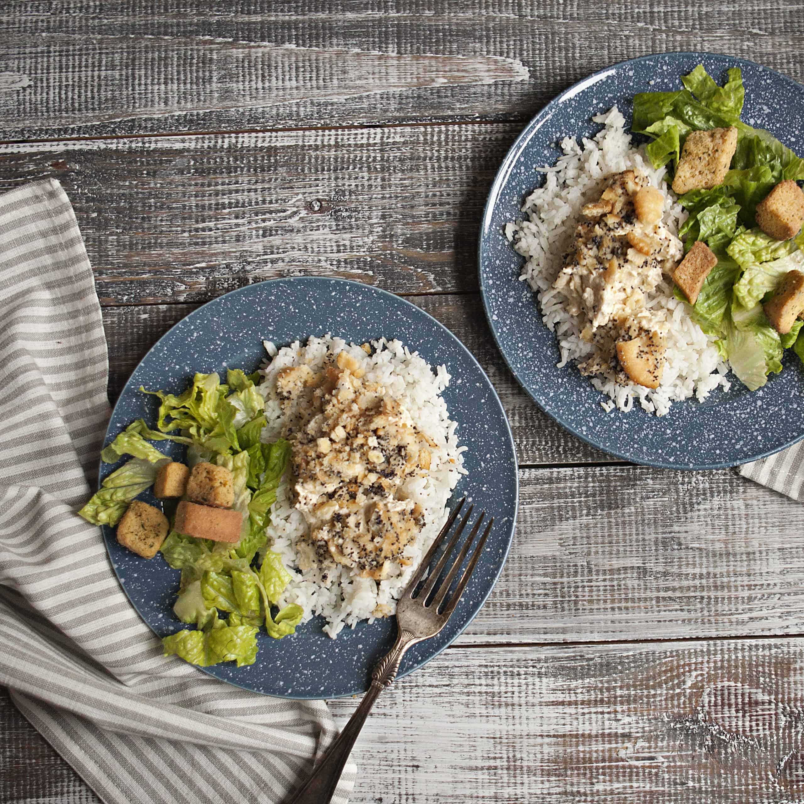 Two blue and white speckled plates containing poppy seed chicken casserole on white rice alongside a green salad with croutons.  One plate has a fork.  The plates are on top of a wooden table next to a gray and white striped kitchen towel.