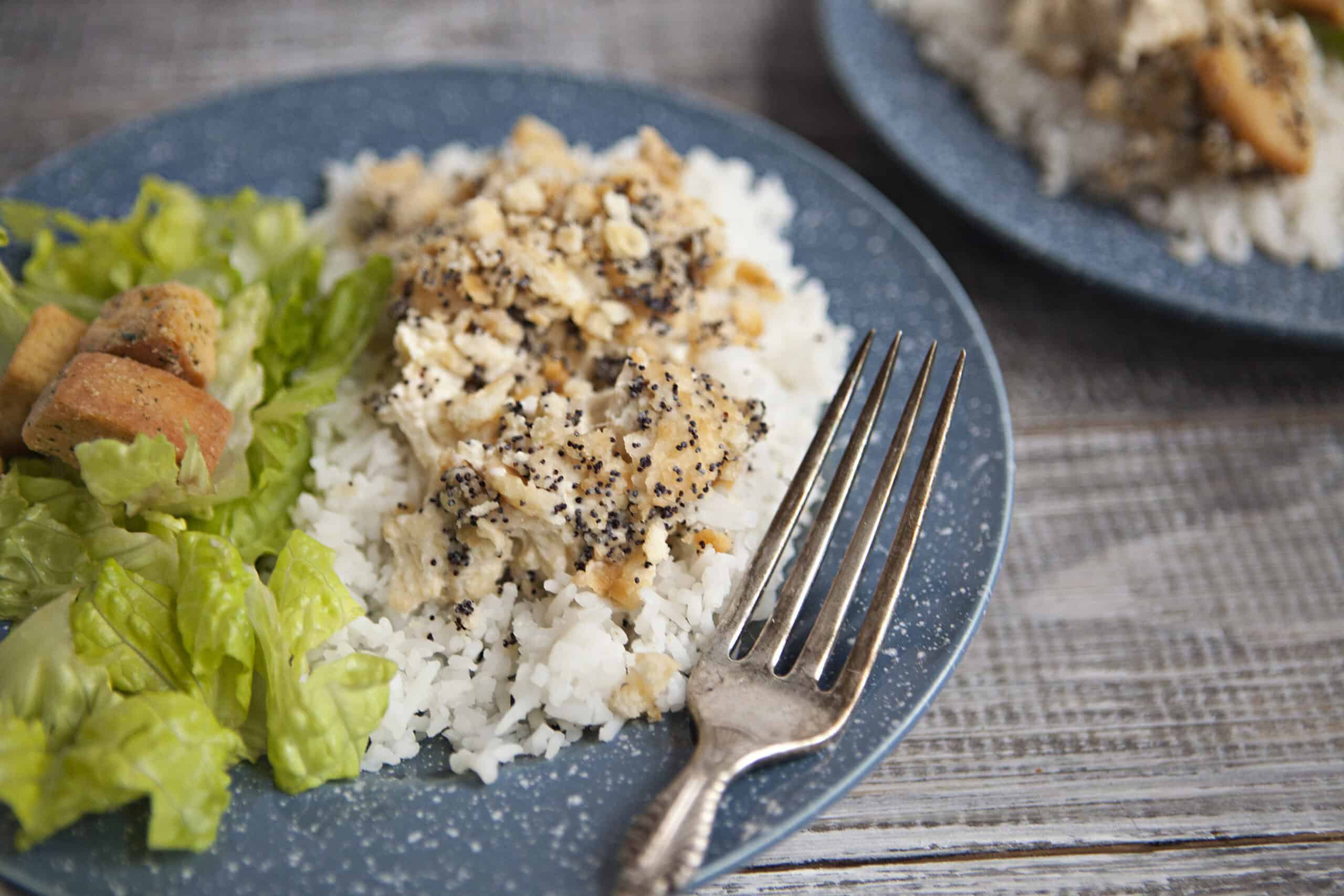 Poppy seed chicken casserole on top of white rice next to a green salad with croutons, alongside a fork, on a wooden table next to a similar plate containing poppy seed chicken and white rice.