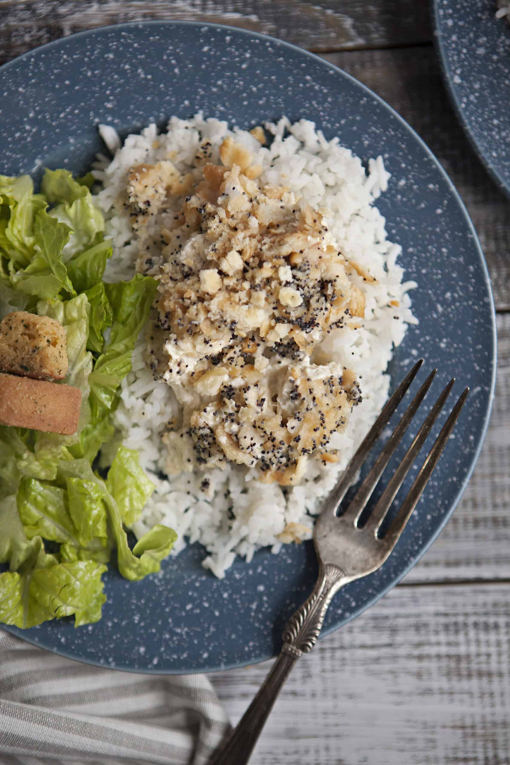 Poppy seed chicken casserole on white rice next to a green salad with croutons on a blue and white speckled plate with a fork, on a wooden table.