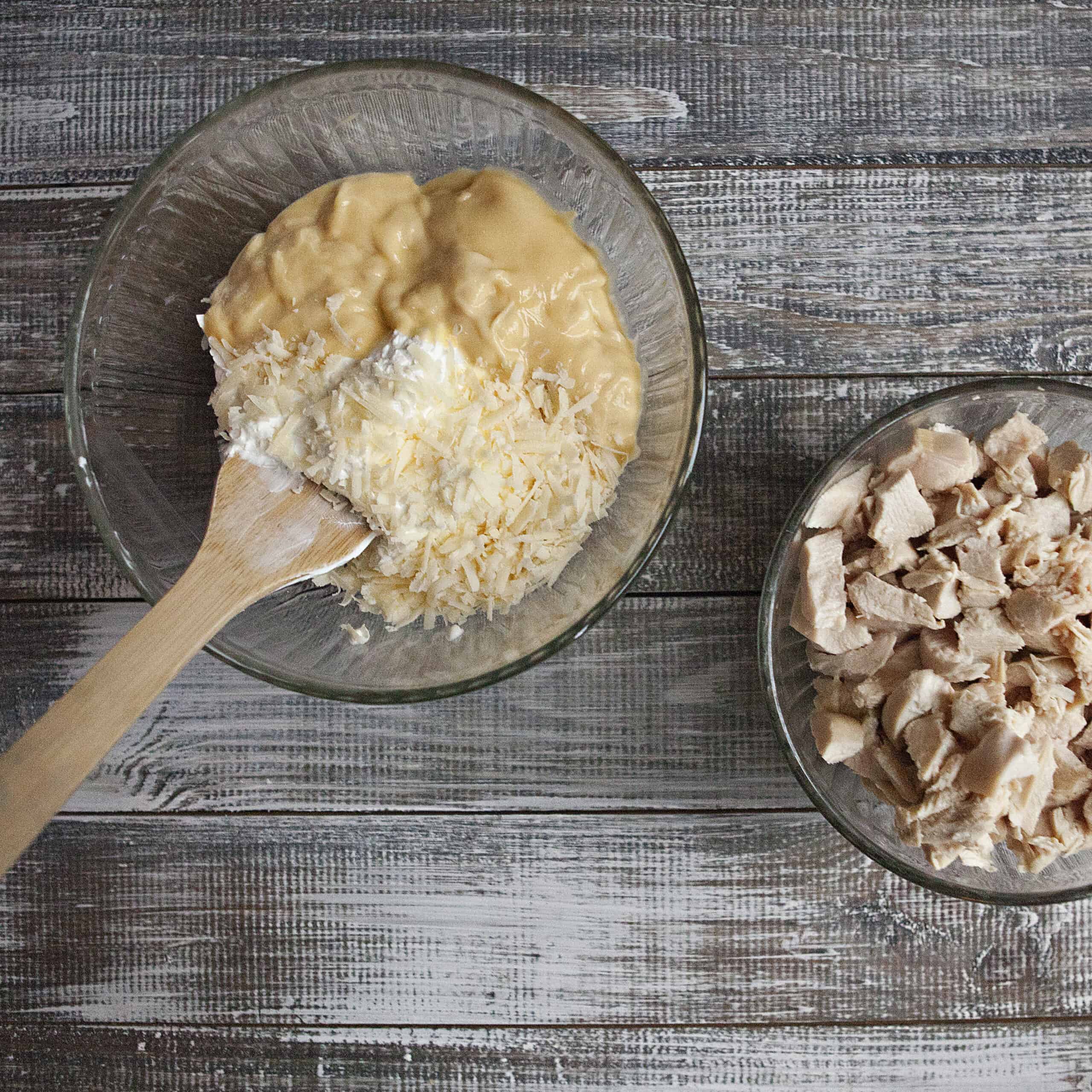 Cream of chicken soup, sour cream, and grated Parmesan cheese in a bowl with a wooden spoon next to another bowl of diced cooked chicken on a wooden table.