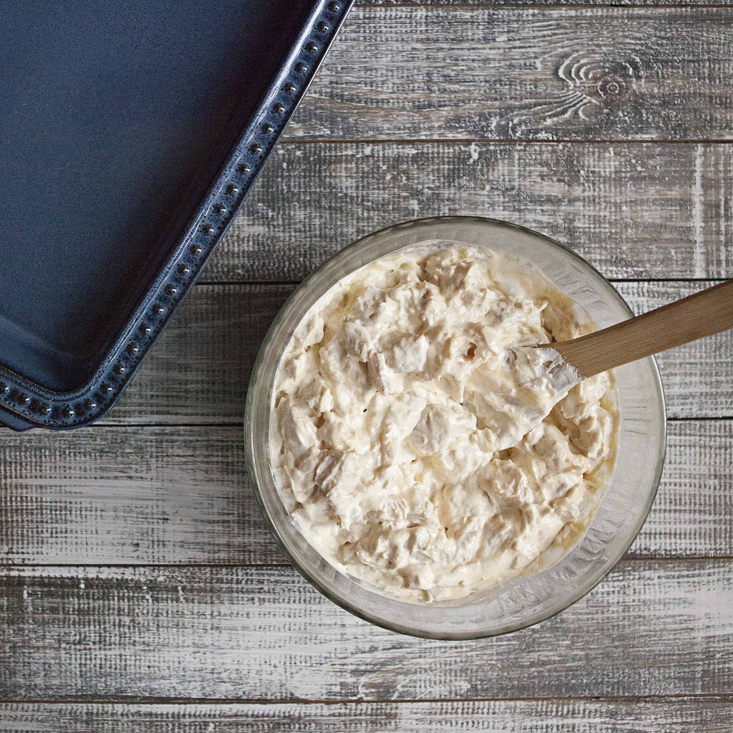 Bowl containing a mixture of diced cooked chicken, cream of chicken soup, sour cream, and grated Parmesan cheese with a wooden spoon next to a blue baking dish on a wooden table.