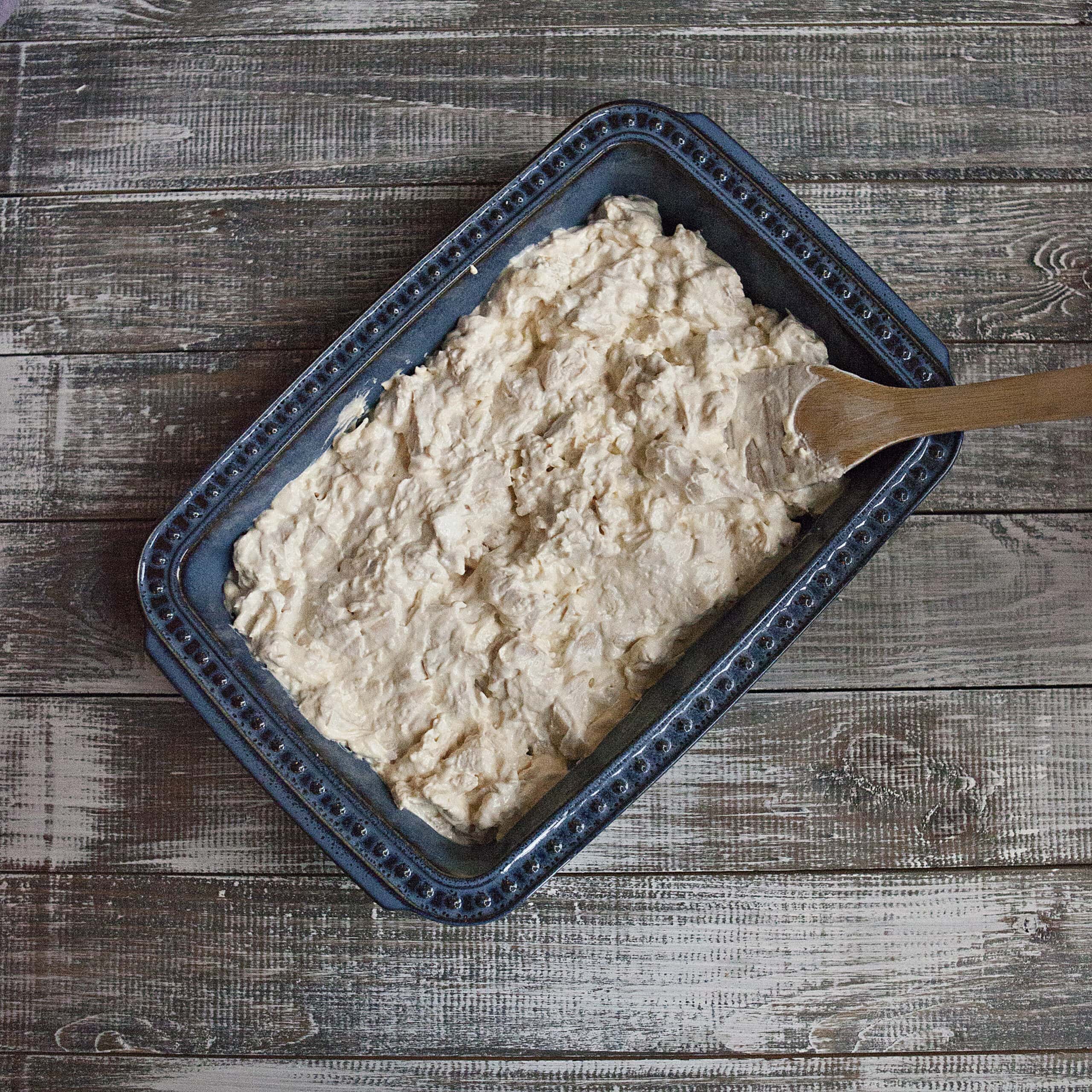 Chicken and sour cream mixture (a precursor to poppy seed chicken casserole) in the bottom of a blue 9x13-inch baking dish on a wooden table.