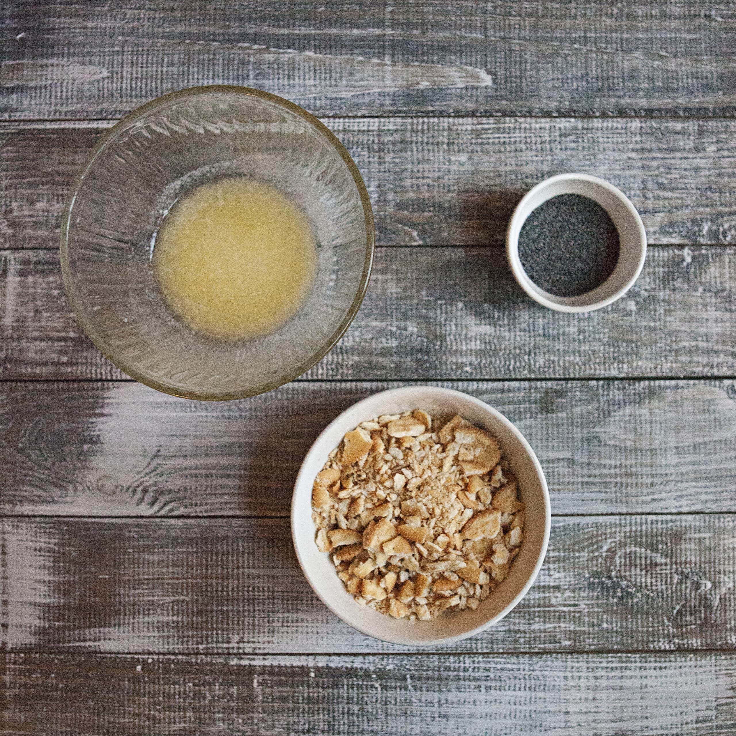 Three bowls containing melted butter, crushed Ritz buttery crackers, and poppy seeds on a wooden table.