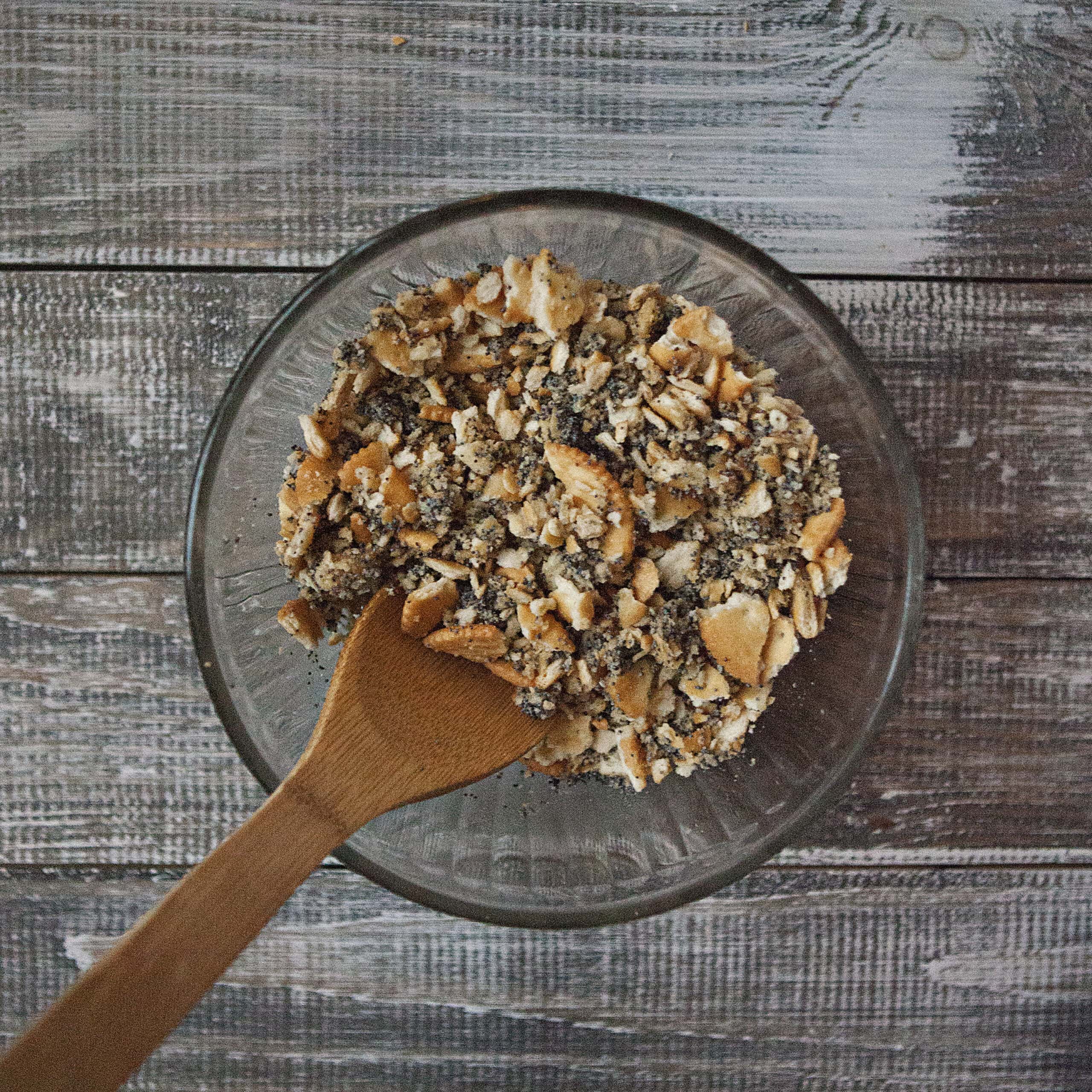 A bowl containing a mixture of crushed Ritz buttery crackers, melted butter, and poppy seeds with a wooden spoon on a wooden table.