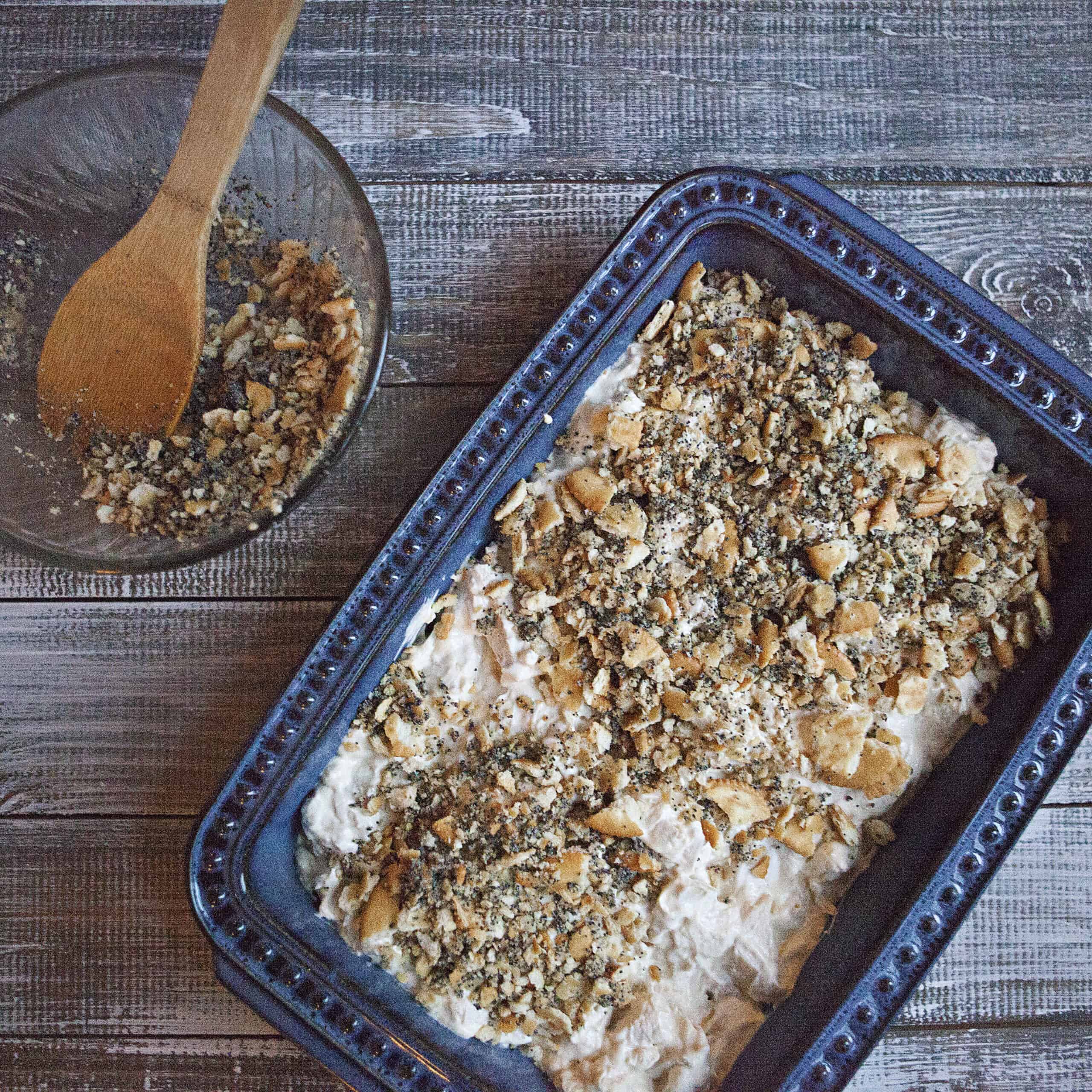 Poppy seed chicken casserole in a blue 9x13-inch baking dish next to a bowl containing more cracker topping for the poppy seed chicken casserole and a wooden spoon on a wooden table.