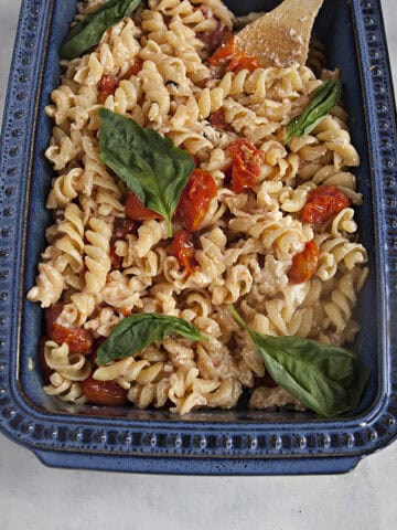 A blue ceramic baking dish filled with baked feta pasta, roasted grape tomatoes, and fresh basil leaves on an off-white background.